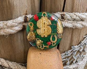 May include: A circular resin charm featuring a green textured base embedded with gold-toned metal charms including a dollar sign, a money bag, a corn cob, a horseshoe, and a key, accented with small red spheres throughout the clear resin surface.
