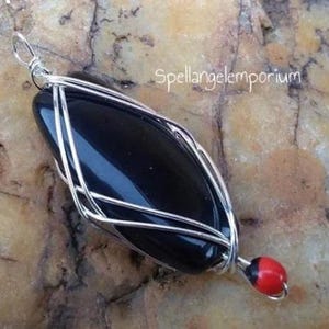 May include: A handcrafted pendant featuring a polished black stone encased in a silver wire cage. A small red and black bead dangles from the bottom. The text "spellangelemporium" is visible in the background.