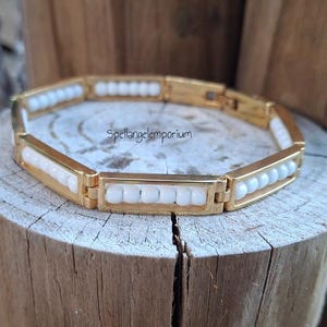 Puede incluir: Pulsera dorada con eslabones rectangulares, cada uno con una fila de cuentas blancas. La pulsera se muestra sobre una superficie de madera. El texto "spellangelemporium" es visible.