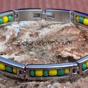 Puede incluir: Un brazalete de acero inoxidable con cuentas verdes y amarillas alternadas. El brazalete está sobre un fondo de roca.