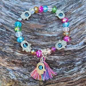 May include: A colorful beaded bracelet with a Hamsa hand charm and a tassel. The bracelet features a variety of faceted beads in shades of blue, pink, yellow, and clear. The Hamsa hand charms are silver-toned.