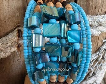 Op de afbeelding: Een wikkelarmband met meerdere strengen turquoise zaadkralen, vierkante iriserende kralen en houten kralen. De armband heeft een bohemien stijl met een mix van texturen en kleuren. De tekst "Spellangelemporium" is zichtbaar.