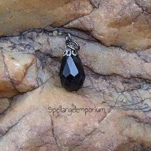 May include: A black teardrop-shaped pendant with a silver bail and a silver crown-like design at the top. The pendant is sitting on a rough, brown rock.