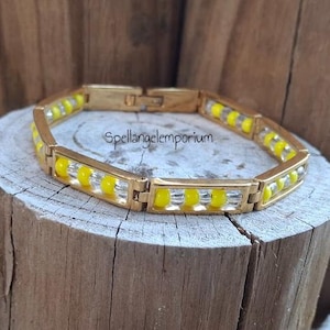 Puede incluir: Pulsera dorada con eslabones rectangulares, cada uno con cuentas amarillas y transparentes. La pulsera se exhibe sobre una superficie de madera. El texto "Spellangelemporium" es visible.