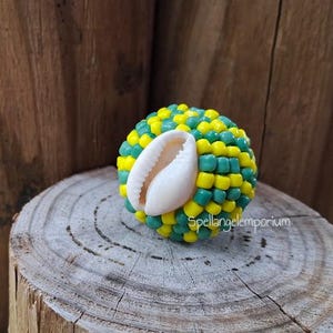 May include: A decorative sphere composed of yellow and teal beads, with a white seashell accent. The sphere is set on a wooden surface, with the text "Spellangelemporium" visible. The background features a wooden texture.