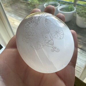 Puede incluir: Una esfera redonda y transparente de selenita con un diseño grabado de carpa koi y nenúfares. La esfera se sostiene en una mano, con luz natural reflejada en su superficie. El diseño es detallado y delicado.