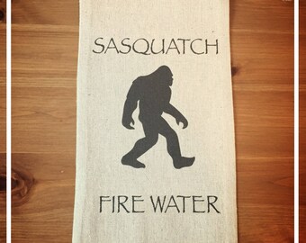 May include: A beige fabric bag with a black silhouette of a Sasquatch and the text "Sasquatch Fire Water".