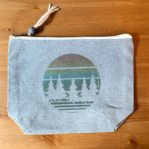 May include: A gray canvas zippered pouch with a white zipper and a white tassel pull. The pouch has a circular design with a rainbow gradient and a silhouette of five pine trees.