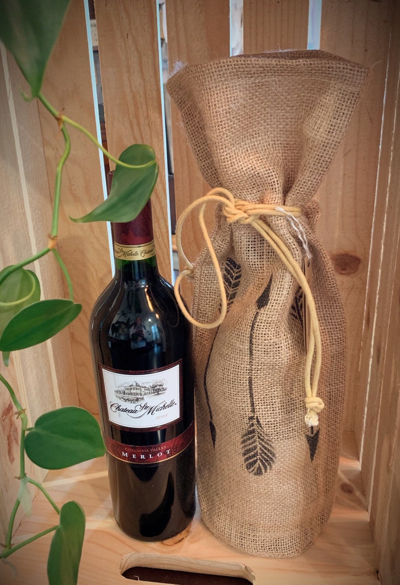 May include: A bottle of Chateau Ste. Michelle Merlot wine in a burlap wine bag with a brown twine tie. The wine bottle is in a wooden crate.