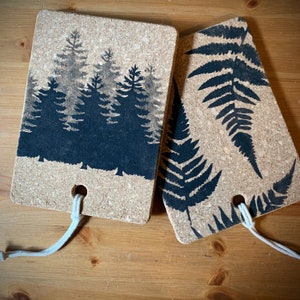 May include: Two rectangular cork coasters. One features a black silhouette of a forest, the other a black fern design. Each has a hole with a white cord for hanging. The coasters rest on a wooden surface.