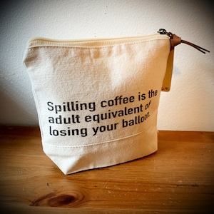May include: A white canvas zippered pouch with a brown leather zipper pull. The pouch has the text "Spilling coffee is the adult equivalent of losing your balloon." printed on it.