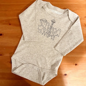 May include: A gray long-sleeve baby onesie with a black line drawing of the Seattle skyline, including the Space Needle and Mount Rainier. The onesie has the text "Shakes + Spreads" on the tag.