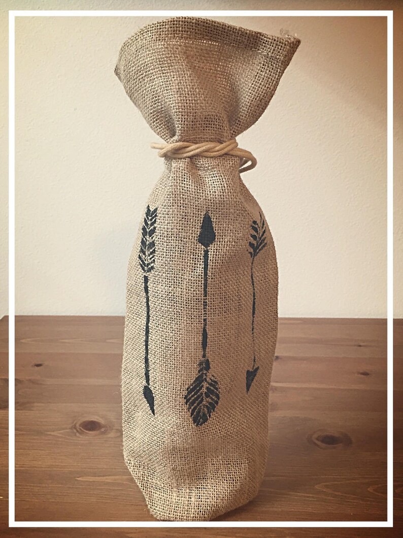 May include: A brown burlap wine bottle bag with a tan rope tie and three black arrow designs.