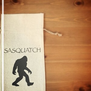 May include: A white canvas bag with the words "Sasquatch Fire Water" printed in black. The bag features a black silhouette of a Sasquatch walking.