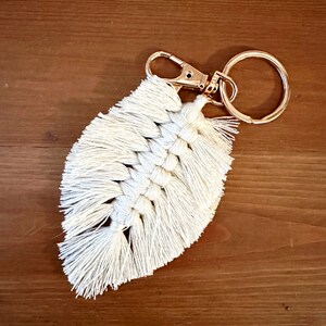 May include: Cream-colored macrame feather keychain with a gold-tone clasp and key ring. The feather design features a woven center and fringed edges, creating a textured and bohemian aesthetic. The keychain is displayed on a wooden surface.