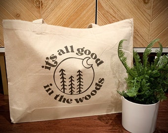 Può includere: Una borsa tote in tela beige con una grafica nera che recita "it's all good in the woods" con una luna crescente e pini stilizzati.