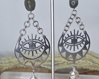 May include: A pair of silver earrings with an eye design. Each earring features a gray stone at the top, connected by a chain to a crescent-shaped pendant with an eye and sun ray design. A faceted clear crystal hangs from the bottom.