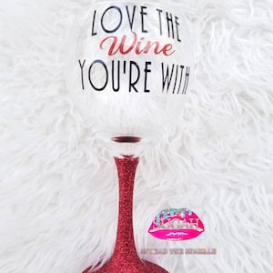 May include: A red glitter wine glass with the text "Love the Wine You're With" on the front. The glass has a pink lip with the text "Sugah" and "Spread the Sparkle" on the bottom.