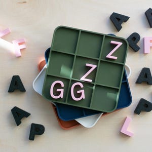 Op de afbeelding: Een groene vierkante bak met een rasterpatroon, gestapeld op andere bakken in wit, blauw en oranje. Roze en zwarte lettertegels liggen verspreid rond de bakken en spellen woorden. De letters zijn ongeveer 2,5 cm hoog.