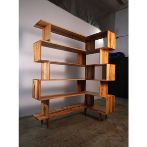 May include: A wooden corner bookshelf with multiple shelves and open compartments. The shelves are made of natural wood with a light brown finish. The bookshelf has a modern design with a geometric shape.