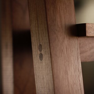 Op de afbeelding: Close-up van een houten plank met twee donkerbruine houten pinnen aan de zijkant van de plank. De plank is gemaakt van massief hout en heeft een natuurlijke afwerking.