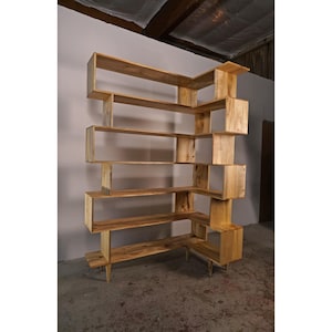 May include: A light-colored wooden bookshelf with an asymmetrical design. The bookshelf has multiple shelves of varying sizes and shapes, creating a unique and modern look. The wood grain is visible, adding to the natural aesthetic. The bookshelf is free-standing and appears to be sturdy.