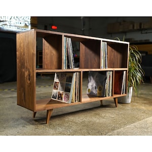 May include: A wooden record shelf with six compartments, each filled with vinyl records. The shelf has a dark brown stain and is sitting on a concrete floor. The shelf has four legs that are slightly angled.