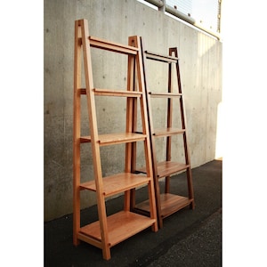 Op de afbeelding: Twee houten ladderplanken, een in licht hout en een in donker hout, tegen een betonnen muur. Beide planken hebben vier planken.