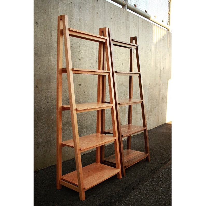 Op de afbeelding: Twee houten ladderplanken, een in licht hout en een in donker hout, tegen een betonnen muur. Beide planken hebben vier planken.