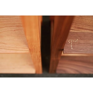 Op de afbeelding: Close-up van een houten plank met twee planken. De bovenste plank is gemaakt van walnoot en heeft een messing inlay in de vorm van een letter 'T'. De onderste plank is gemaakt van kersenhout.