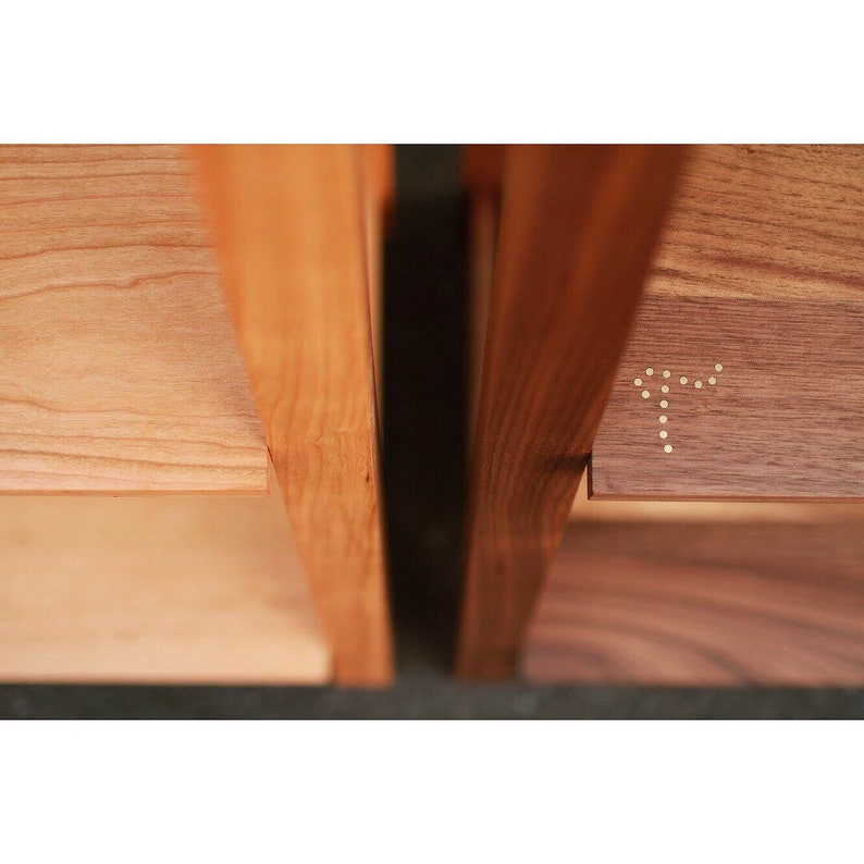 Op de afbeelding: Close-up van een houten plank met twee planken. De bovenste plank is gemaakt van walnoot en heeft een messing inlay in de vorm van een letter 'T'. De onderste plank is gemaakt van kersenhout.