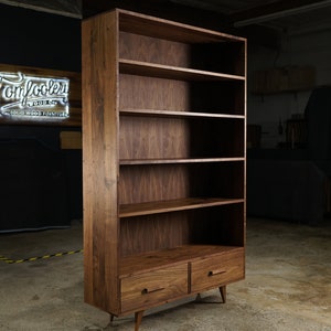 May include: A tall, solid wood bookcase with a warm brown finish. It features multiple shelves and two drawers at the bottom. The bookcase has a mid-century modern design with tapered legs and simple drawer pulls.