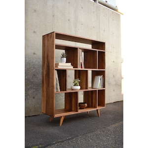 May include: A wooden bookcase with 16 open shelves, featuring a mid-century modern design with tapered legs. The bookcase is filled with books, plants, and decorative items.
