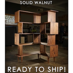 May include: A solid walnut bookshelf with an open, geometric design. The shelving unit features multiple cubby spaces and a unique, angular shape. The text "SOLID WALNUT" and "READY TO SHIP!" are displayed in white.
