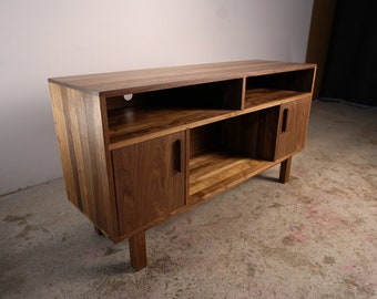 Könnte beinhalten: Eine dunkelbraune Holz-TV-Konsole mit zwei Türen und offenen Regalen. Die Konsole hat ein modernes Design mit klaren Linien und einer natürlichen Holzoptik.