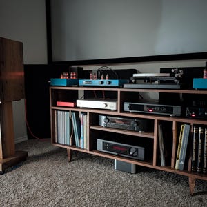 K&ouml;nnte beinhalten: Ein h&ouml;lzernes Audiosystem mit einem gro&szlig;en Lautsprecher auf einem St&auml;nder und einem Schrank im Mid-Century-Modern-Stil. Der Schrank enth&auml;lt verschiedene Audiokomponenten, Schallplatten und B&uuml;cher. Der Lautsprecher ist etwa 102 cm hoch.