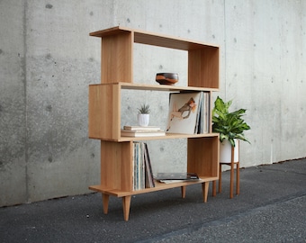 Könnte beinhalten: Ein Bücherregal aus hellem Holz mit vier Regalbrettern und einem einzigartigen Design. Die Regalbretter sind versetzt übereinander gestapelt und das Bücherregal hat vier Beine. Das Bücherregal ist mit Büchern, Schallplatten und einer kleinen Topfpflanze gefüllt.