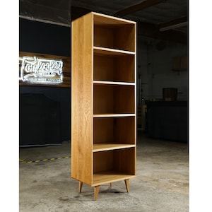 May include: A five-shelf bookcase made of light brown wood with a simple design. The bookcase has four legs that are slightly angled outward. The bookcase is standing on a concrete floor.