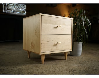 May include: A light wood nightstand with two drawers and gold-toned handles. The nightstand has four legs and is sitting on a concrete floor.