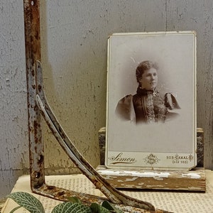 May include: A rusty white metal shelf bracket with a curved design. The bracket is resting on a stack of old books with a vintage photograph of a woman in a dark dress on top. The photograph has the text "Simon 929 CANAL ST (old 183)" printed on the bottom.