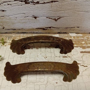May include: Two antique, rusty metal drawer pulls with ornate, scalloped edges. The pulls are a dark brown colour and have a distressed finish.
