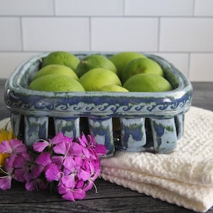May include: A blue and green ceramic fruit basket with a wavy design around the rim. The basket is filled with limes and is sitting on a white towel on a wooden surface.