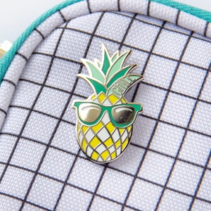 May include: A white and black grid patterned zippered pouch with a turquoise zipper pull. A pineapple with sunglasses enamel pin is attached to the pouch.