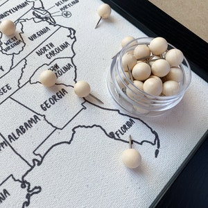 May include: A close-up of a map of the United States with wooden push pins marking different locations. The map is framed and has a white background with black lines outlining the states. The push pins are in a small clear container.