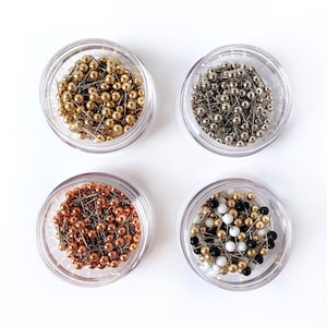 May include: Four small clear containers filled with colorful push pins. The containers are filled with gold, silver, copper, and black and white push pins.