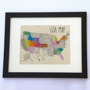 May include: A framed print of a hand-drawn map of the United States. Each state is outlined and filled with a different color, and the state name is written inside the outline. The map is titled "USA Map" in black text.