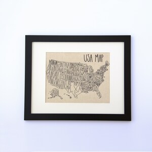 May include: A black framed print of a USA map with the names of each state written inside the outline of the state. The map is on a beige canvas background. The title "USA MAP" is written at the top of the map.