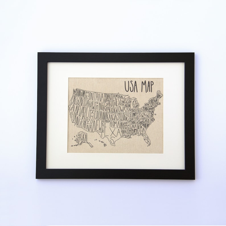 May include: A black framed print of a USA map with the names of each state written inside the outline of the state. The map is on a beige canvas background. The title "USA MAP" is written at the top of the map.
