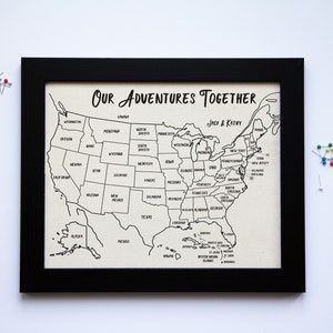 May include: A black framed map of the United States with the text "Our Adventures Together" and "Jack & Katy" printed on it. The map is divided into states and is ready for push pins.