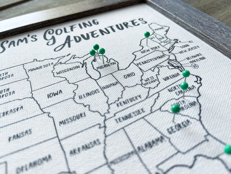 Op de afbeelding: Een witte canvaskaart van de Verenigde Staten met groene duimspijkers die verschillende locaties markeren. De kaart heeft de titel "Sam's Golfing Adventures".